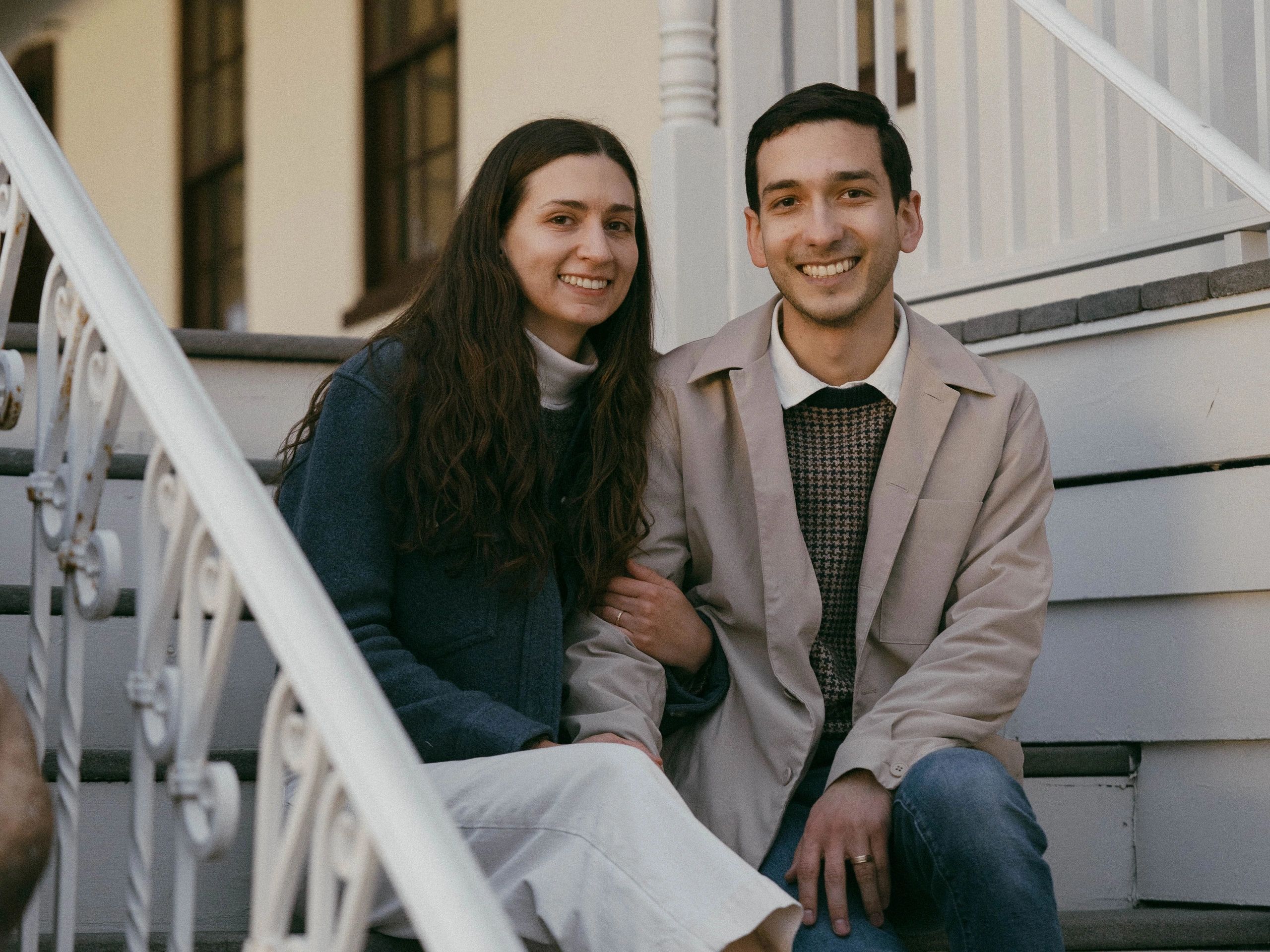 Smiling couple sitting on outdoor stairs, holding hands and looking at the camera.